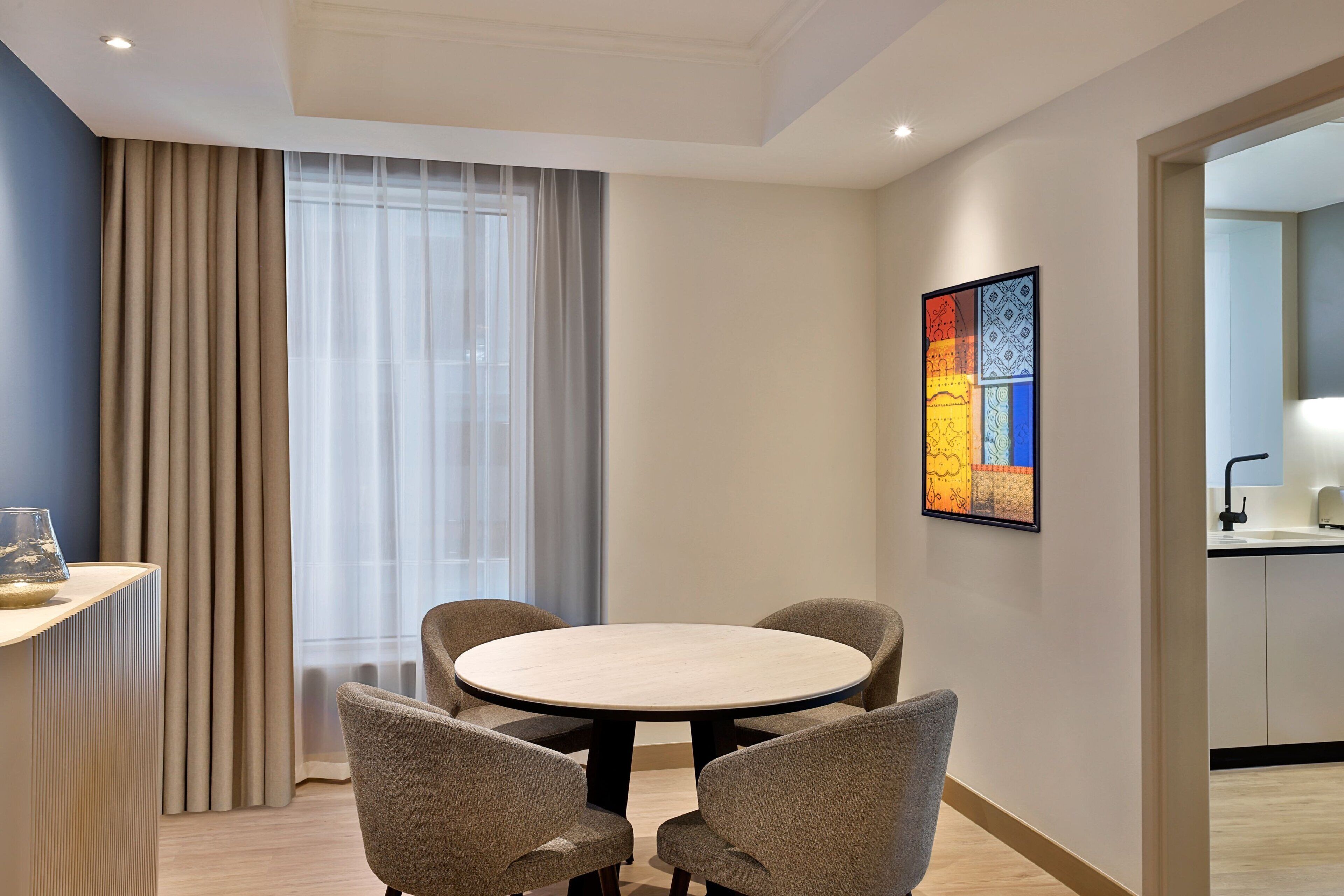 A modern hotel room with a round dining table, four chairs, a large window with curtains, a kitchen area, and a colorful artwork on the wall.