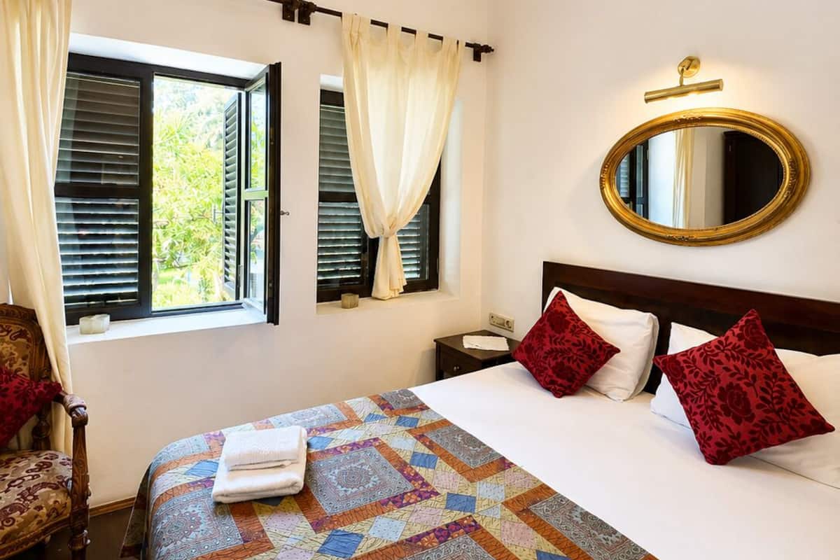 A hotel room with a large bed, a chandelier, a window with shutters, a mirror, and a wooden chair.