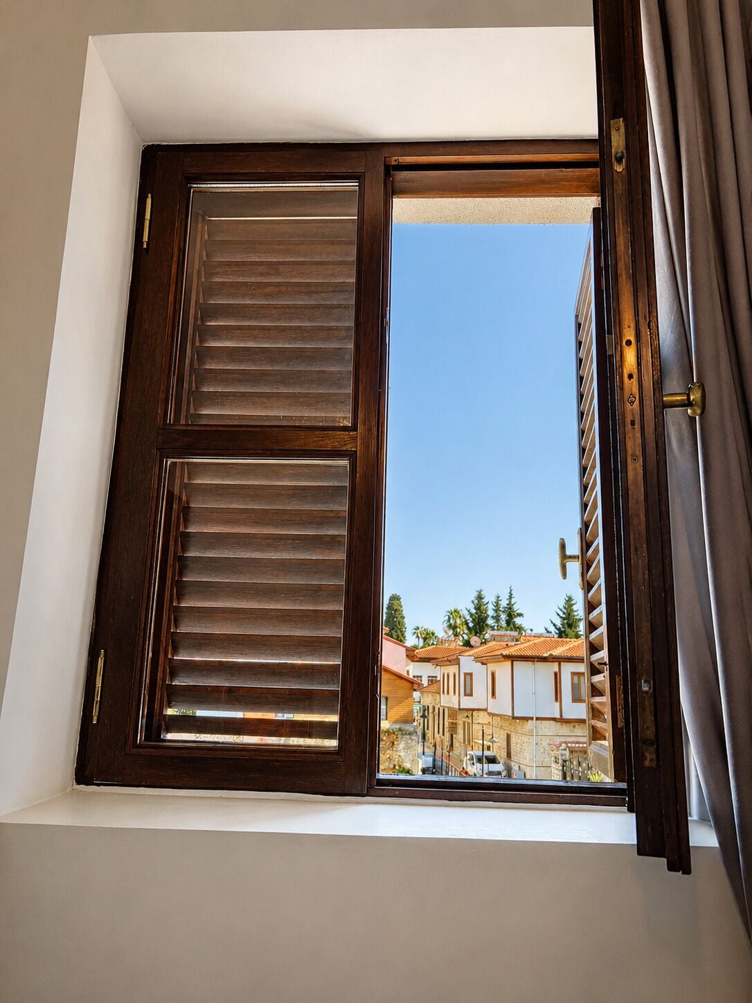 A window with wooden shutters, a view of buildings, and a clear sky.