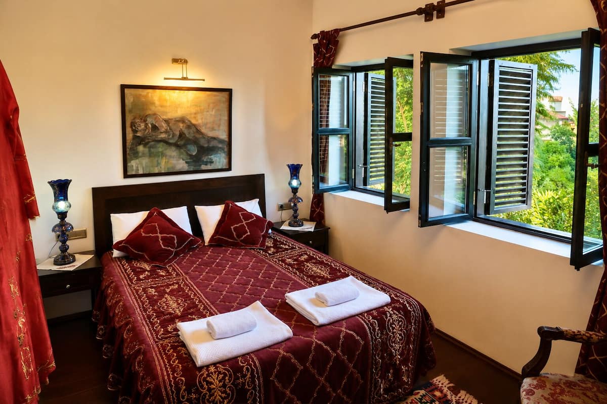 A neatly made bed with a red bedspread, two white towels, and a painting above the headboard.