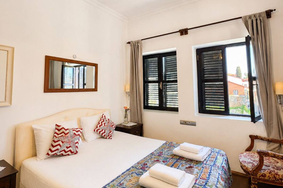 A bedroom with a large bed, a chandelier, a mirror, two windows with shutters, and a chair.