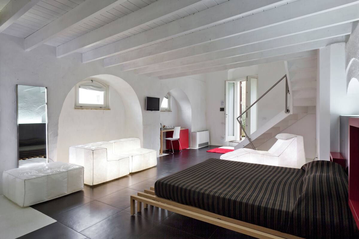 A modern hotel room with a large bed, white furniture, a red accent wall, and a staircase.