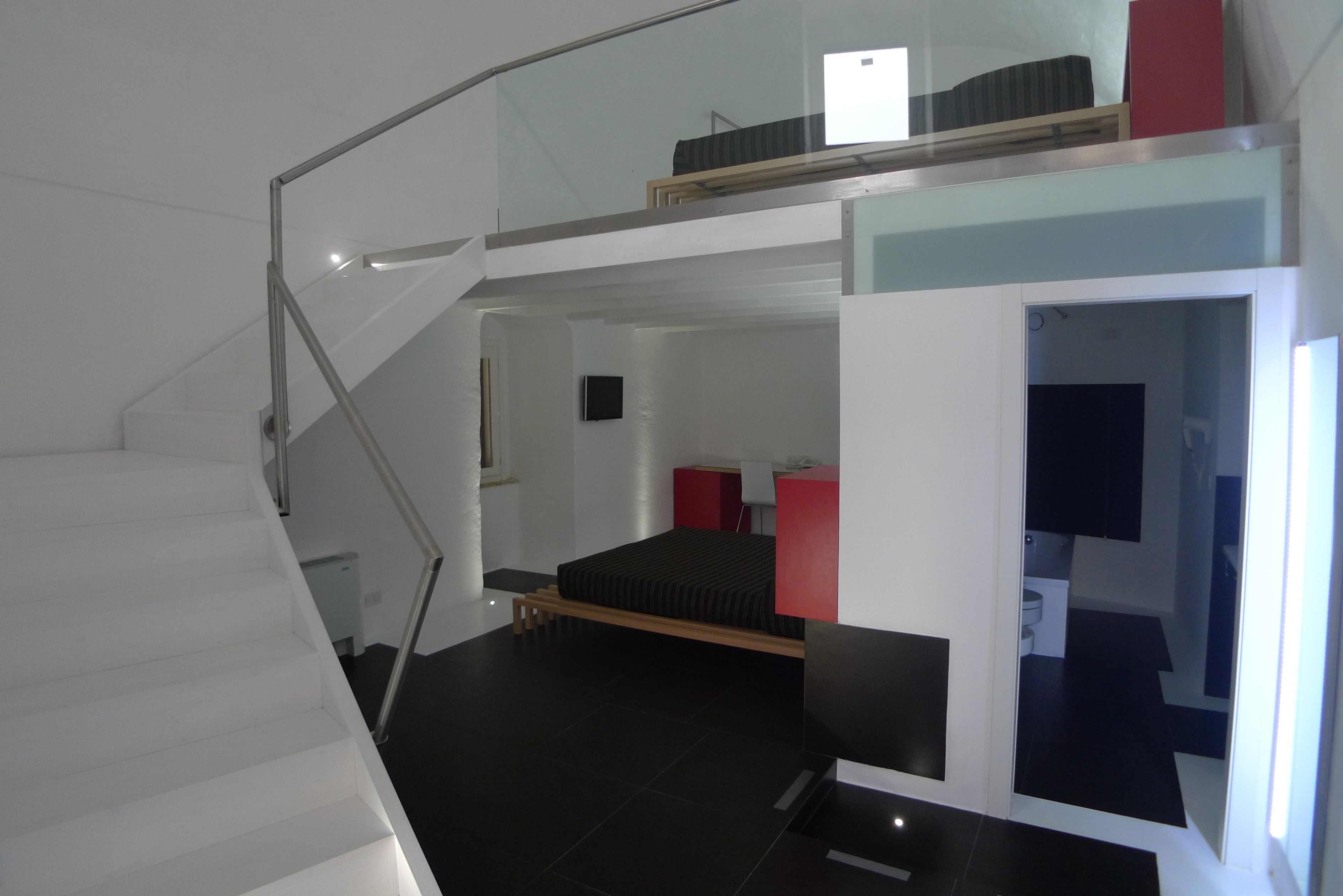 A modern hotel room with a staircase, a bed, a small desk, and a bathroom visible through an open door.