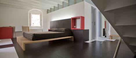 A modern bedroom with a bed, a red accent wall, and a staircase.