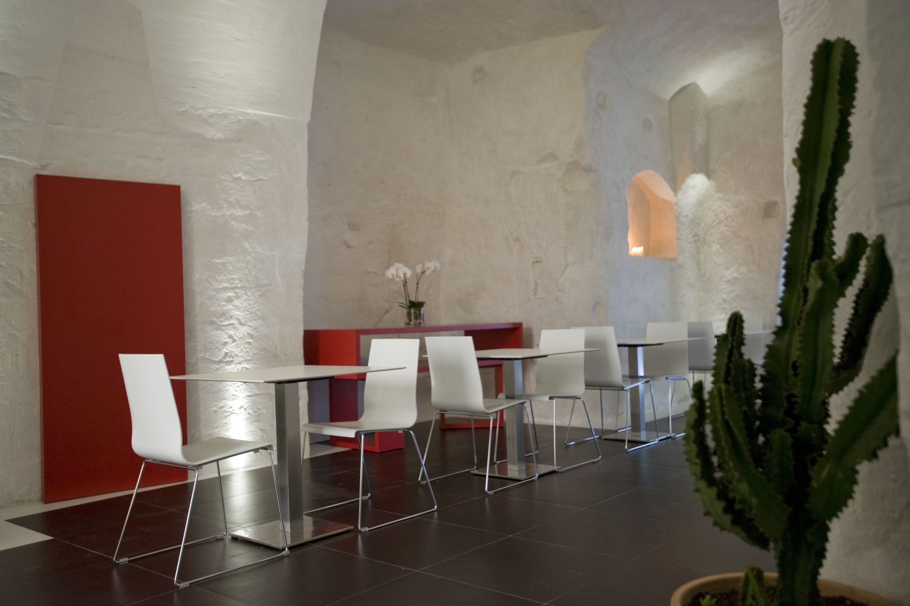 Een moderne eethoek met witte stoelen en een glazen tafel, een rode accentmuur en een cactussplant.