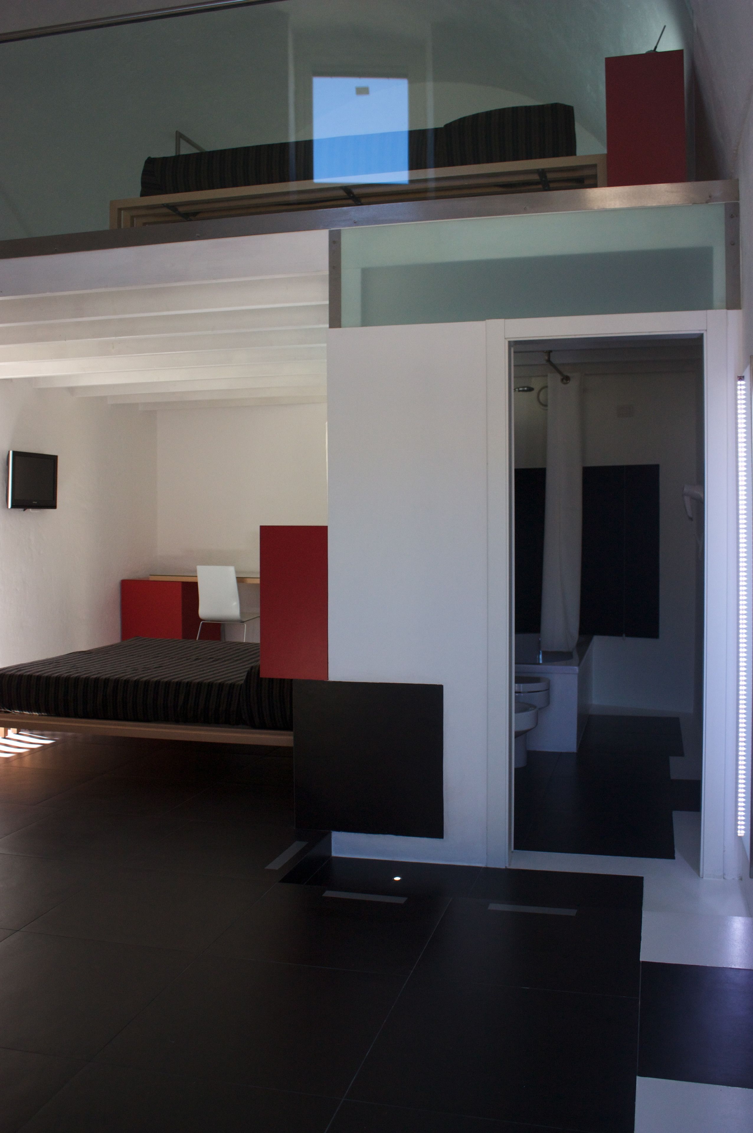 A modern room with a bed, a desk, and a chair. There is a bathroom visible through an open door.
