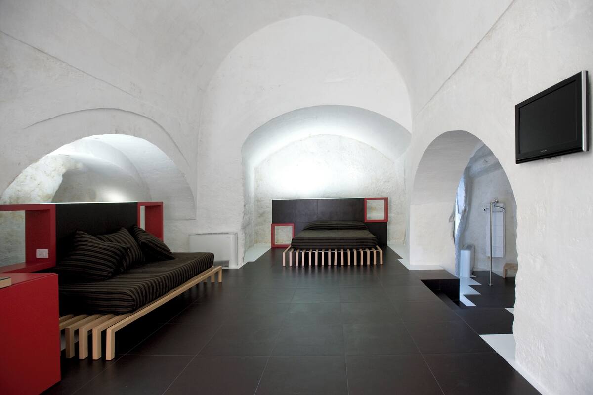A modern hotel room with a minimalist design, featuring a bed, a red seating area, and a television mounted on the wall.