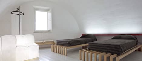 A minimalist bedroom with two wooden beds, a white sofa, and a window providing natural light.