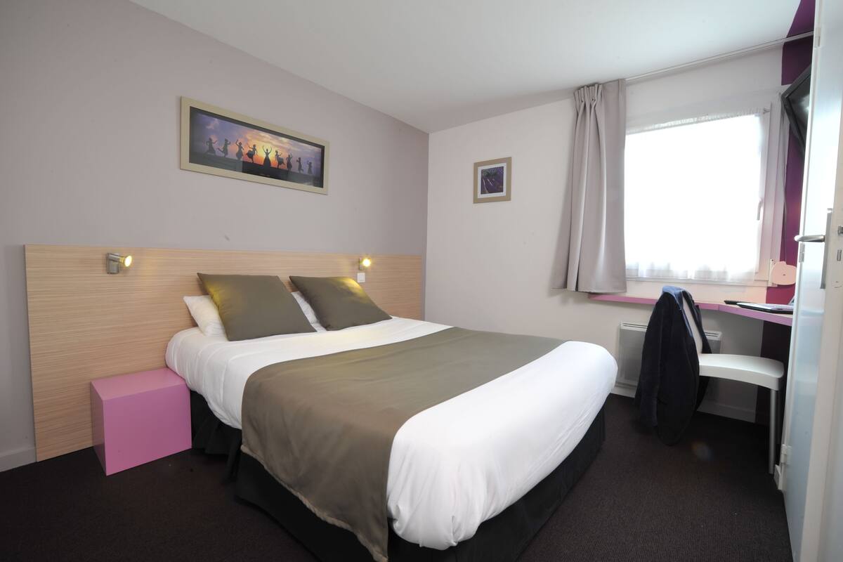 Ein Hotelzimmer mit einem Bett, einem Schreibtisch, einem Stuhl, einer kleinen rosafarbenen Schachtel und einem Fenster mit Vorhängen.