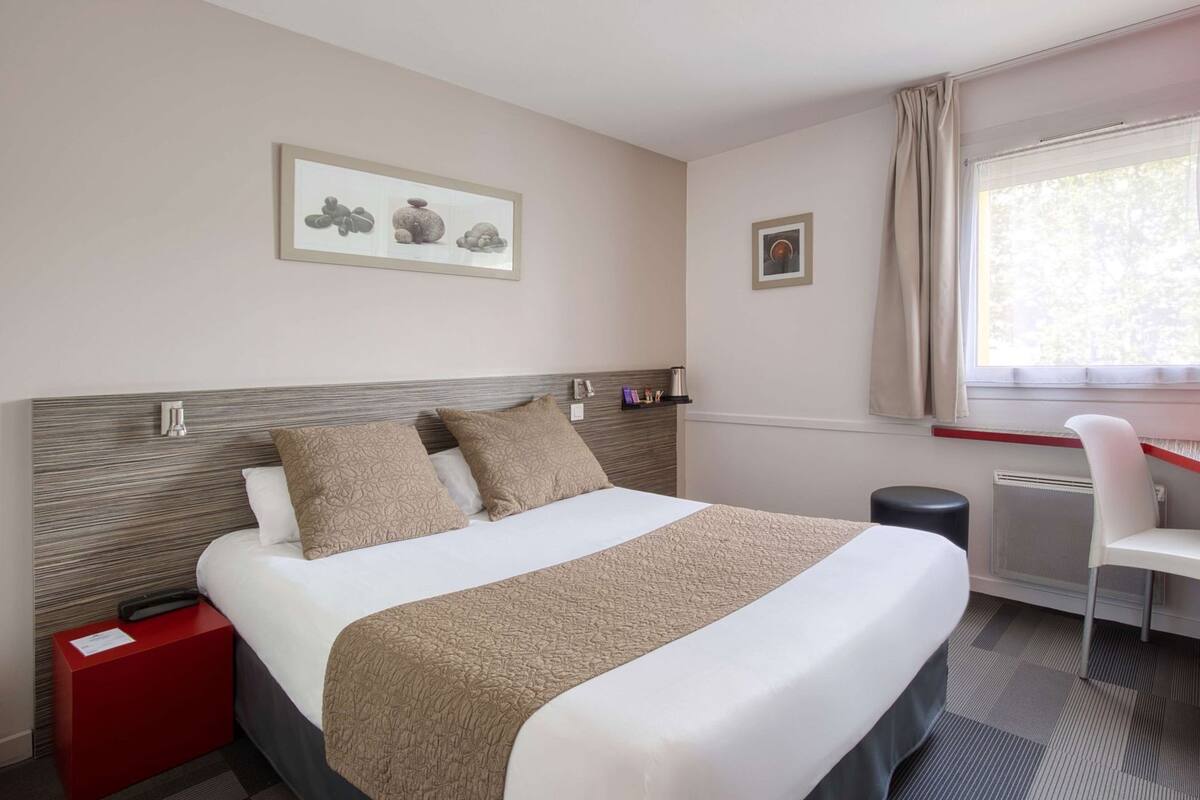 Ein Hotelzimmer mit Bett, Schreibtisch, Stuhl, einem roten Nachttisch und einem Fenster mit Vorhängen.