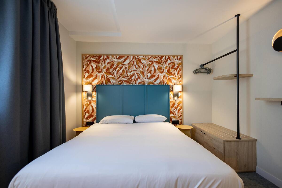 Ein ordentlich eingerichtetes Hotelzimmer mit einem großen Bett, Nachttischlampen, einem Holznachttisch und einem Wandregal.