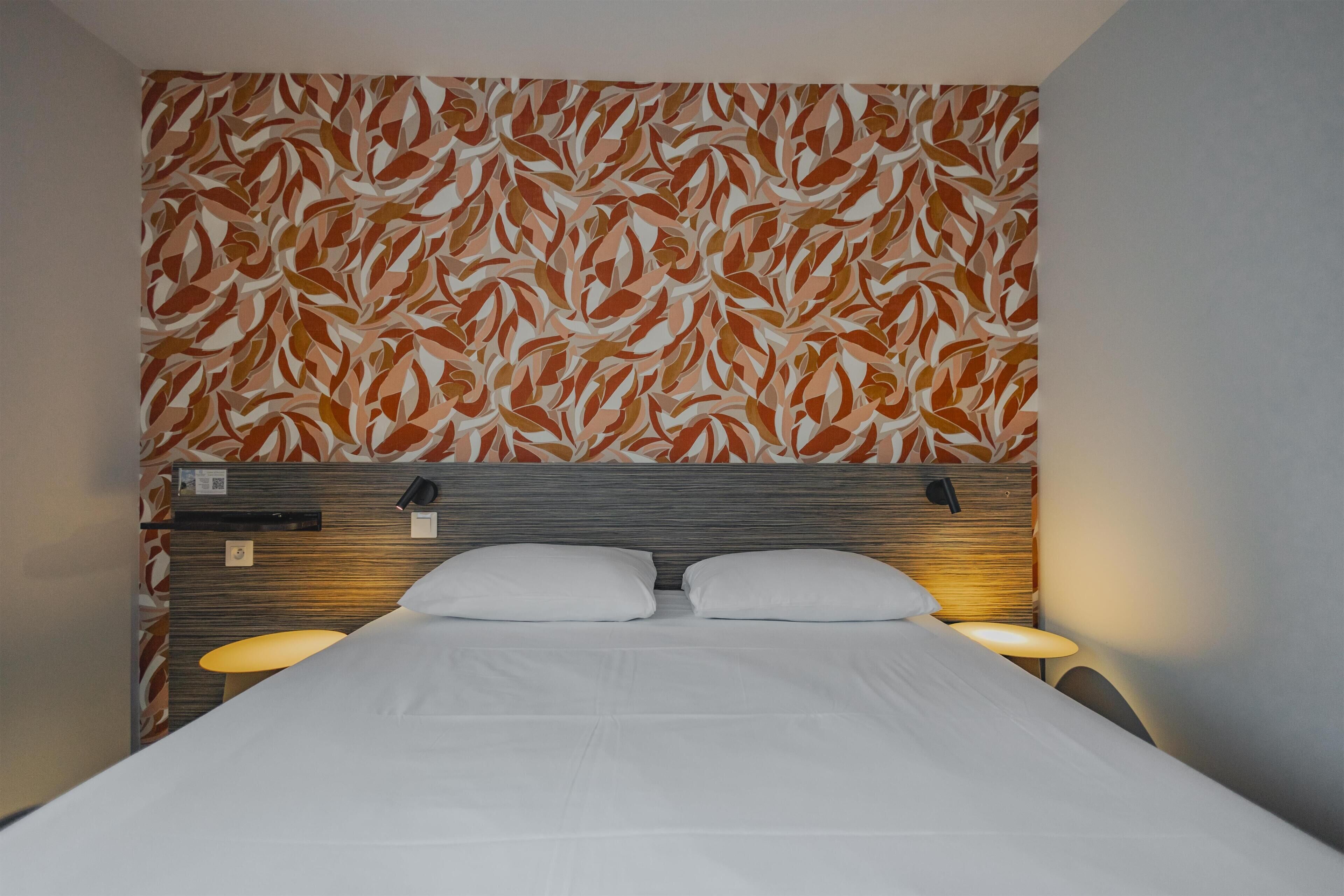 A hotel room with a bed, two bedside lamps, a headboard, and a patterned wall.