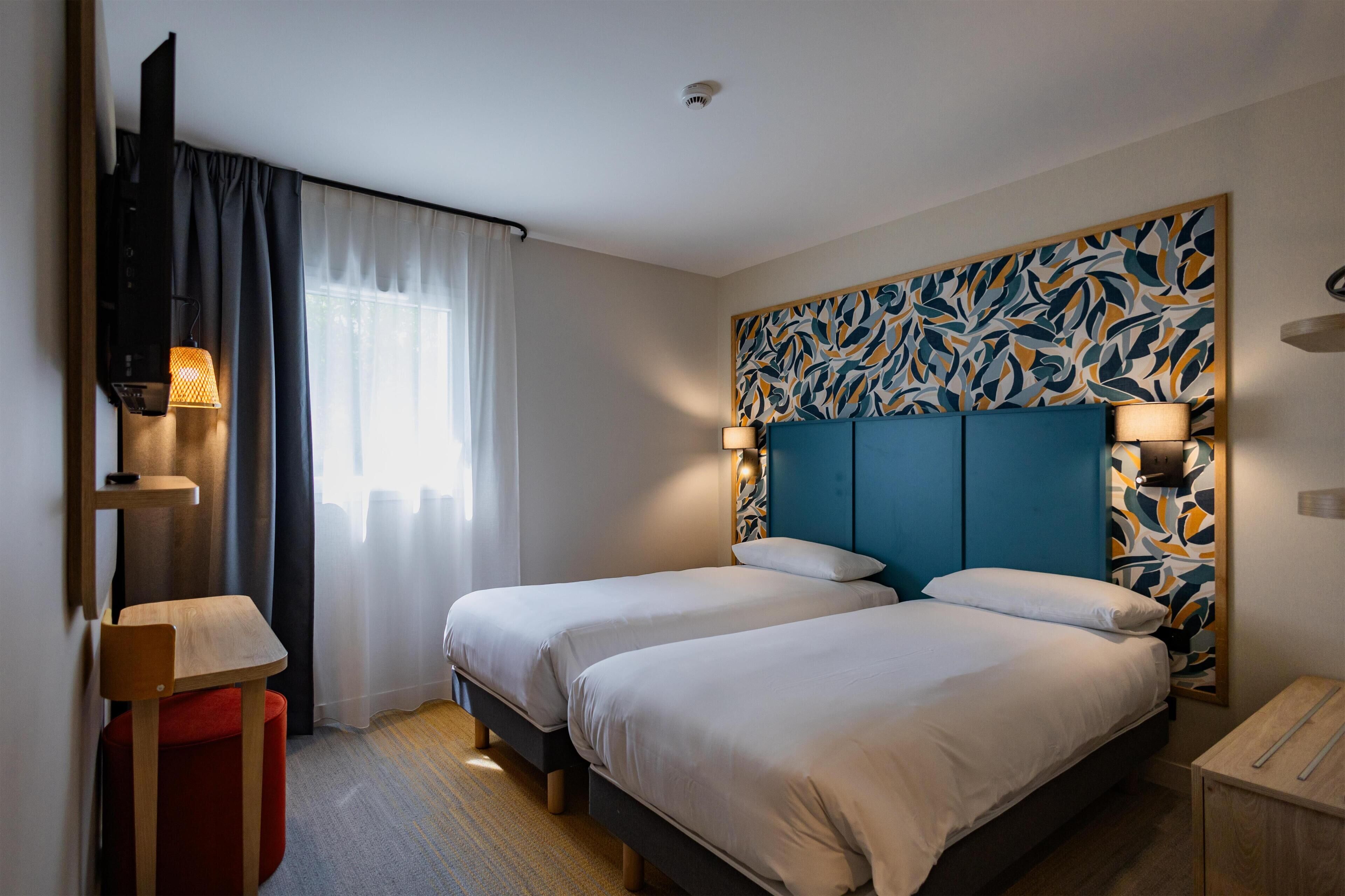 A hotel room with two beds, a TV, a desk, and a red ottoman.