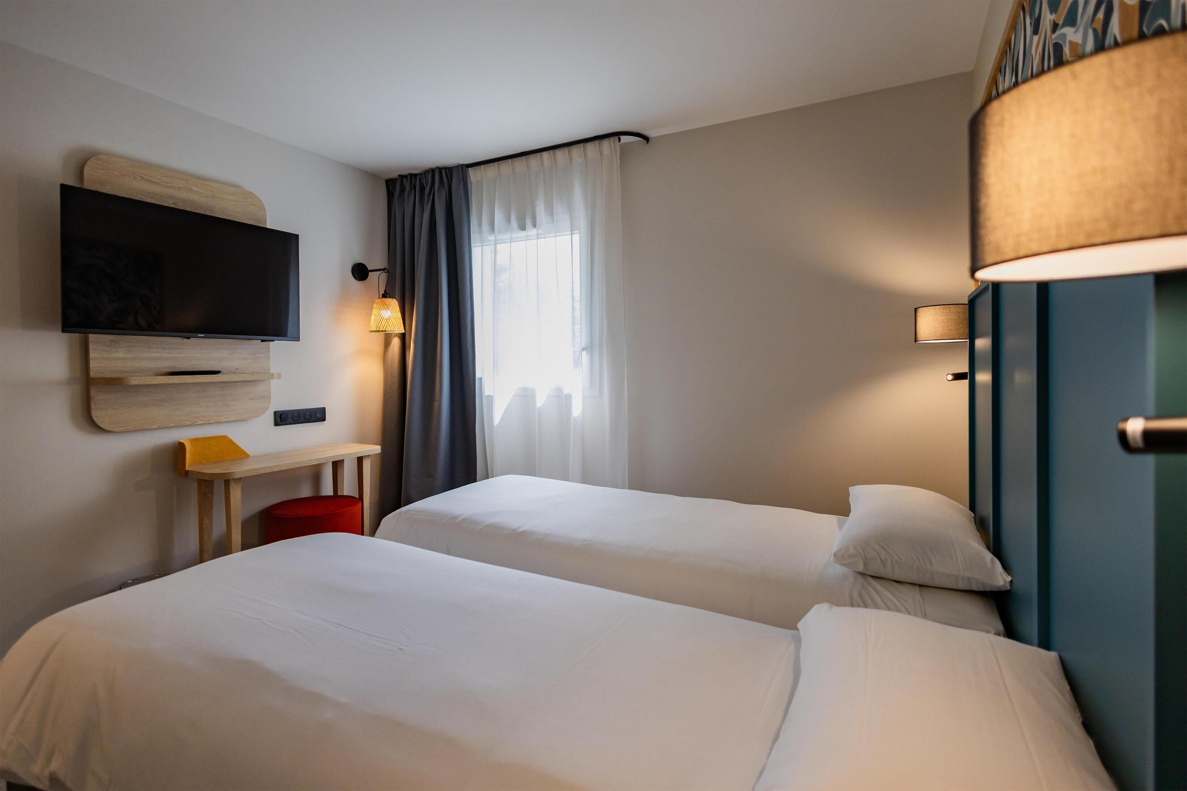 Habitación de hotel con dos camas, un televisor de pantalla plana montado en la pared, una mesita y una lámpara.