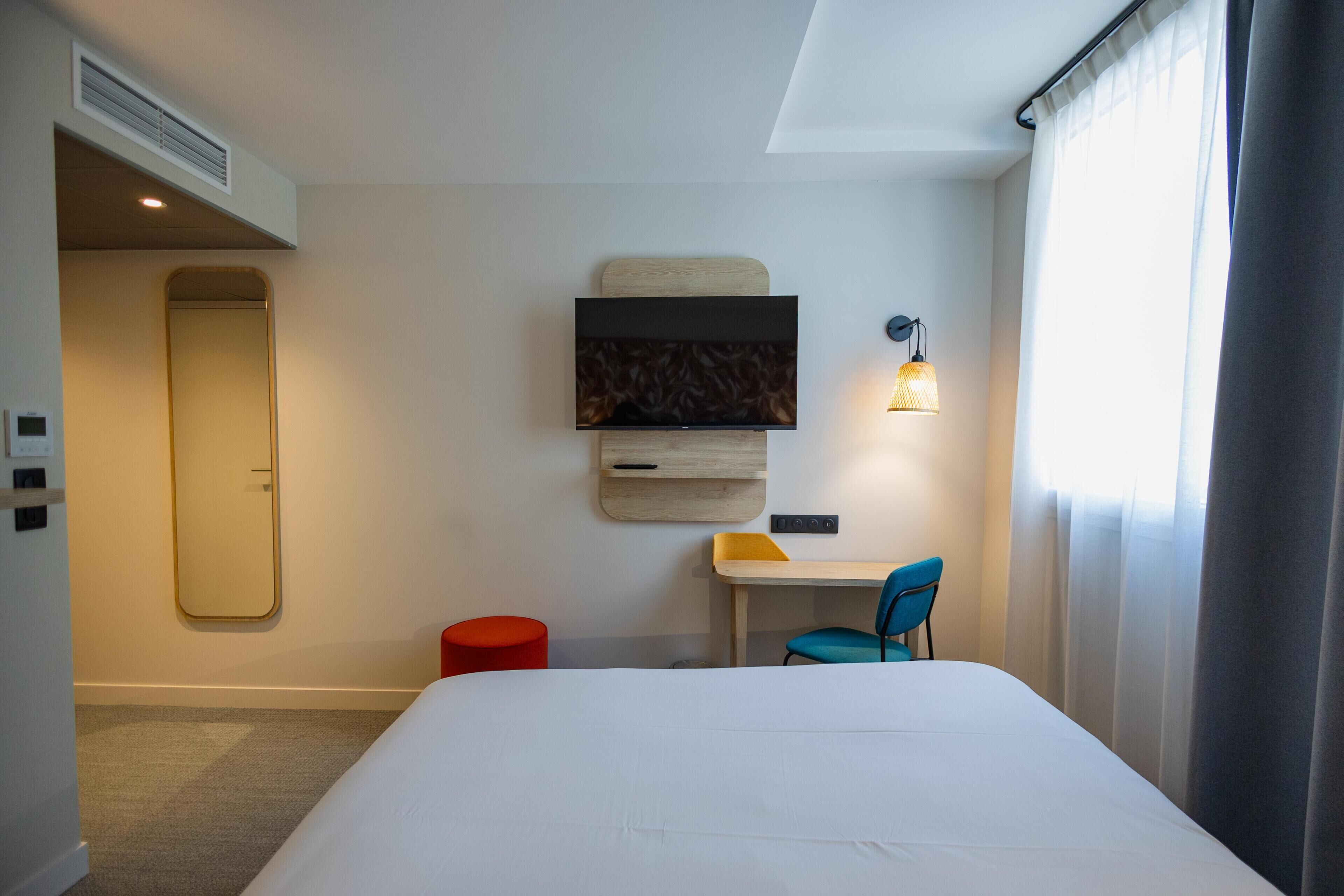 Una habitación de hotel con cama, escritorio, silla, espejo, televisor de pantalla plana y lámpara de pared.