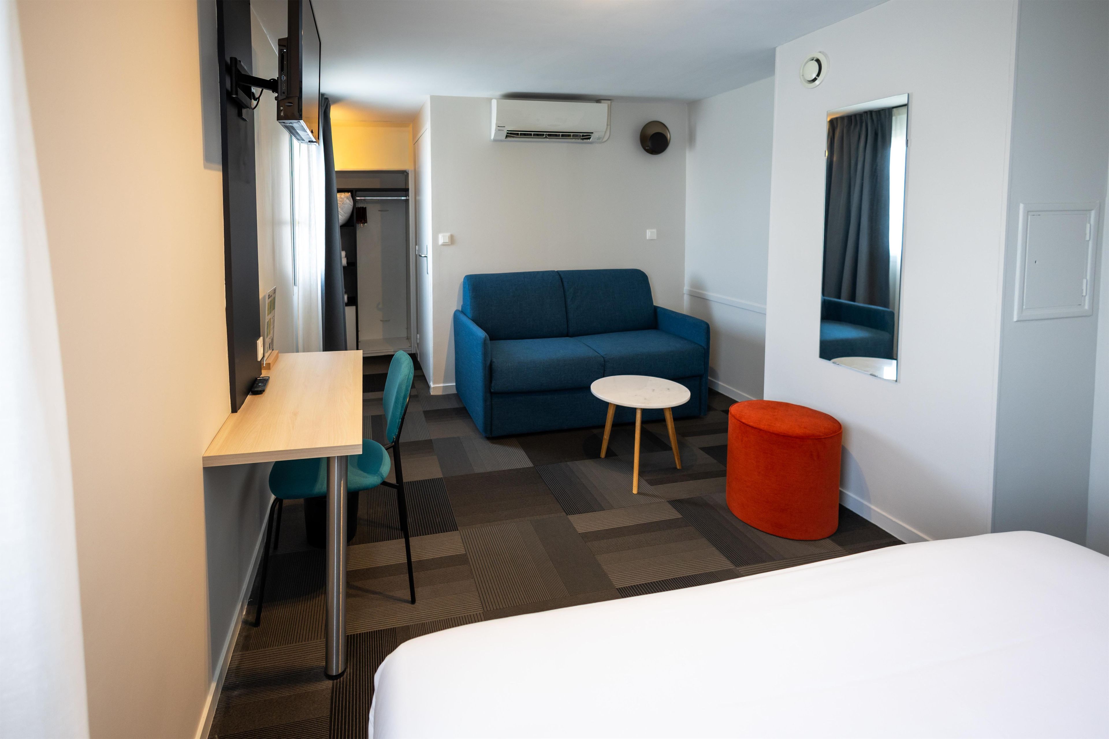 A hotel room with a bed, a desk, a blue sofa, a small round table, and a red ottoman.
