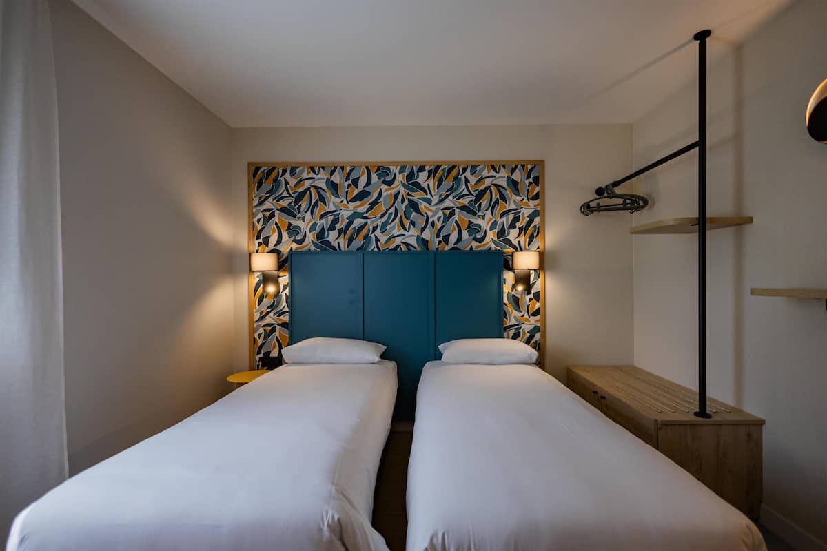 Ein Hotelzimmer mit zwei Einzelbetten, einem blauen Kopfteil, Nachttischlampen, einem Holznachttisch und einem an der Wand montierten Regal.