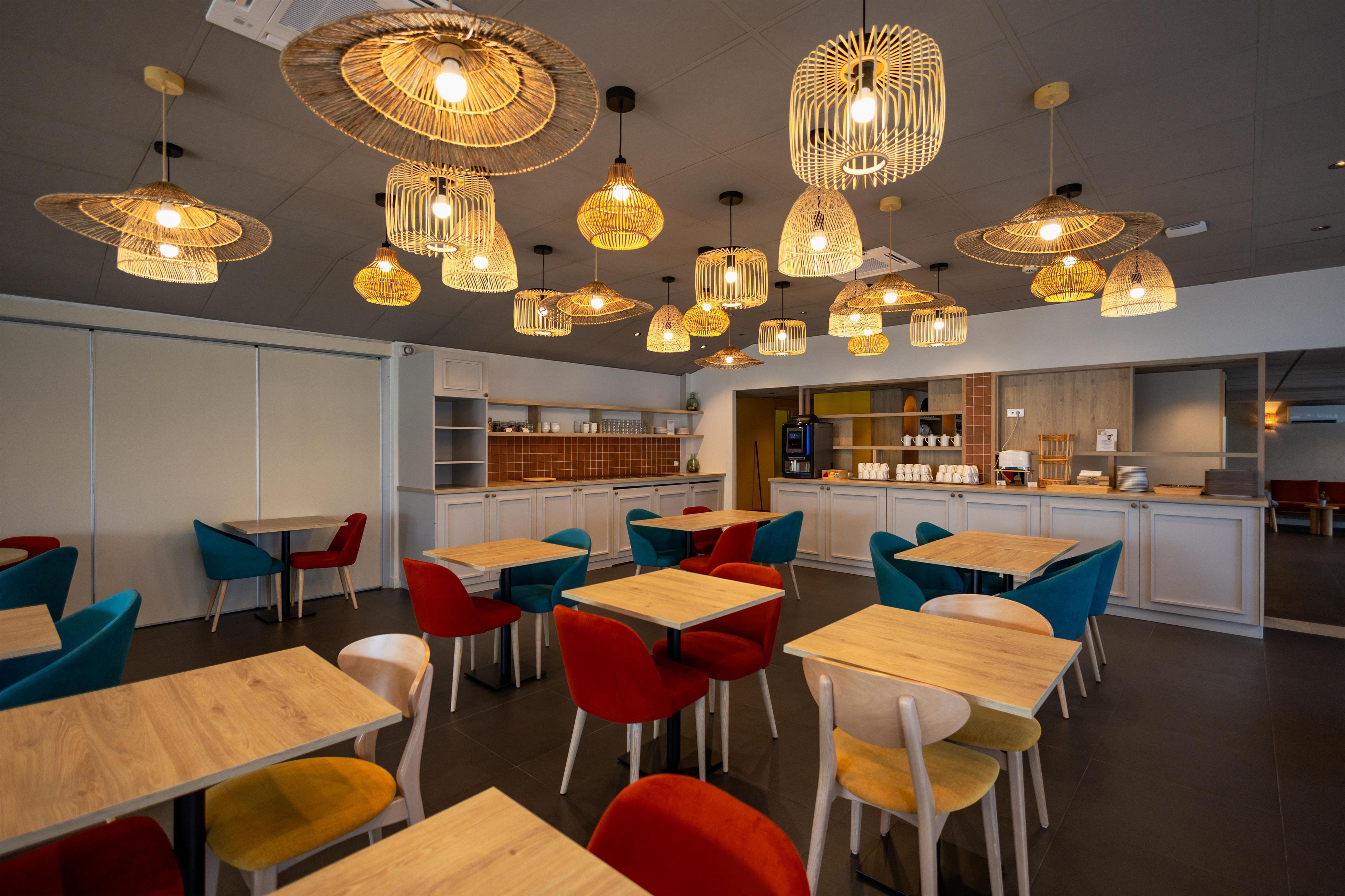 A modern restaurant interior with multiple dining tables and chairs, a kitchen area, and hanging pendant lights.