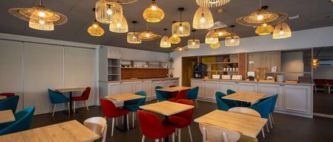 A modern restaurant interior with multiple dining tables and chairs, a kitchen area, and hanging pendant lights.