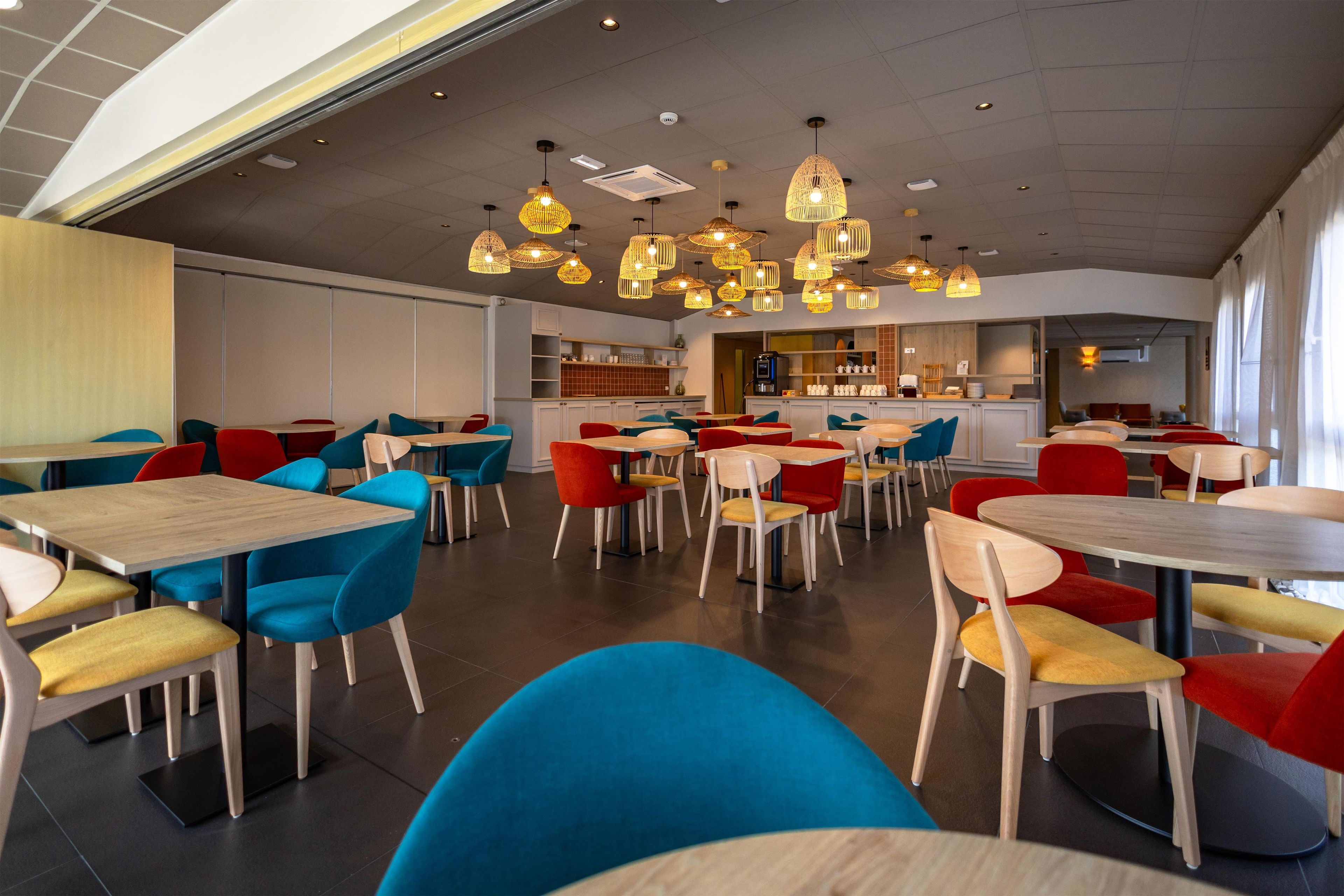 A modern restaurant interior with multiple dining tables and chairs, overhead lighting, and a kitchen area.