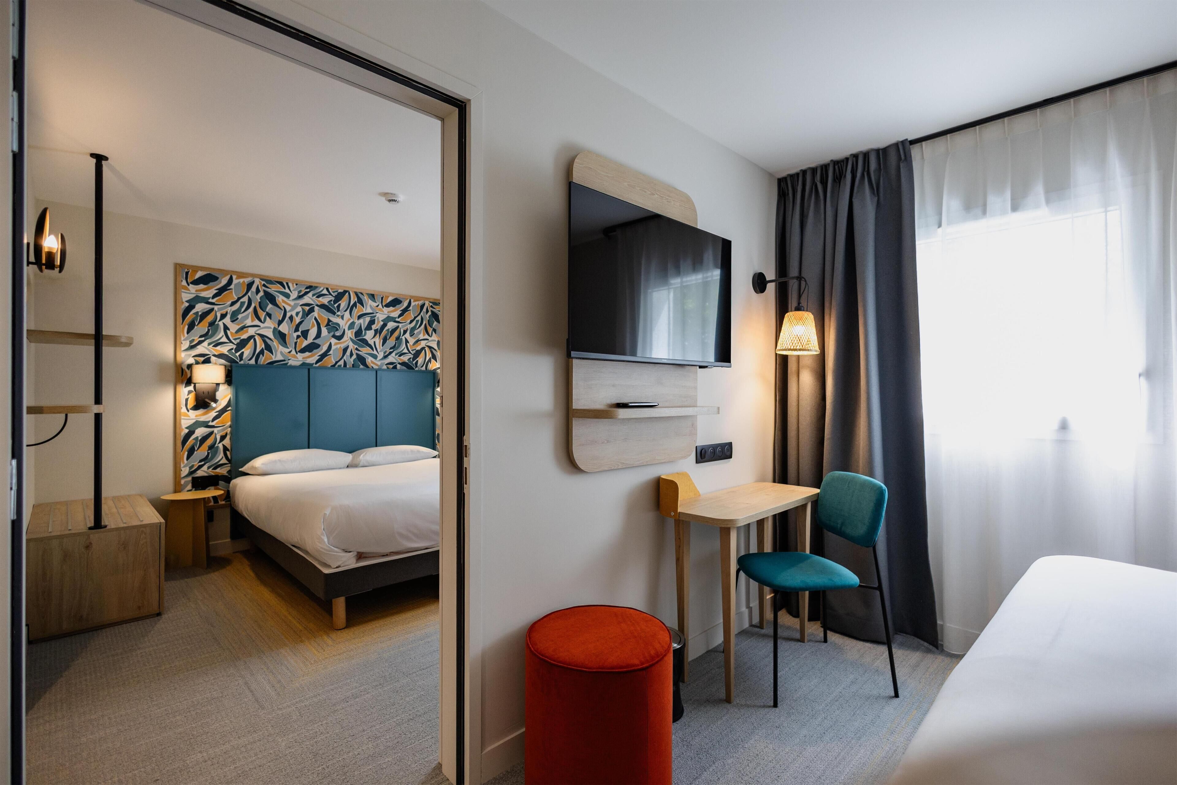 Una habitación de hotel con cama, escritorio, silla, televisor y espejo.
