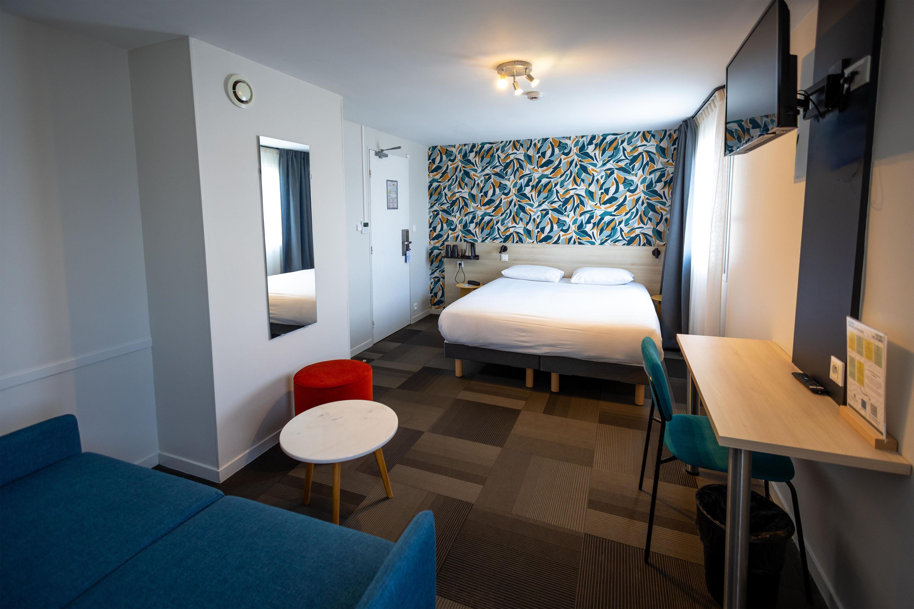 A hotel room with a bed, a desk, a chair, a small table, a red ottoman, a mirror, and a patterned wall.