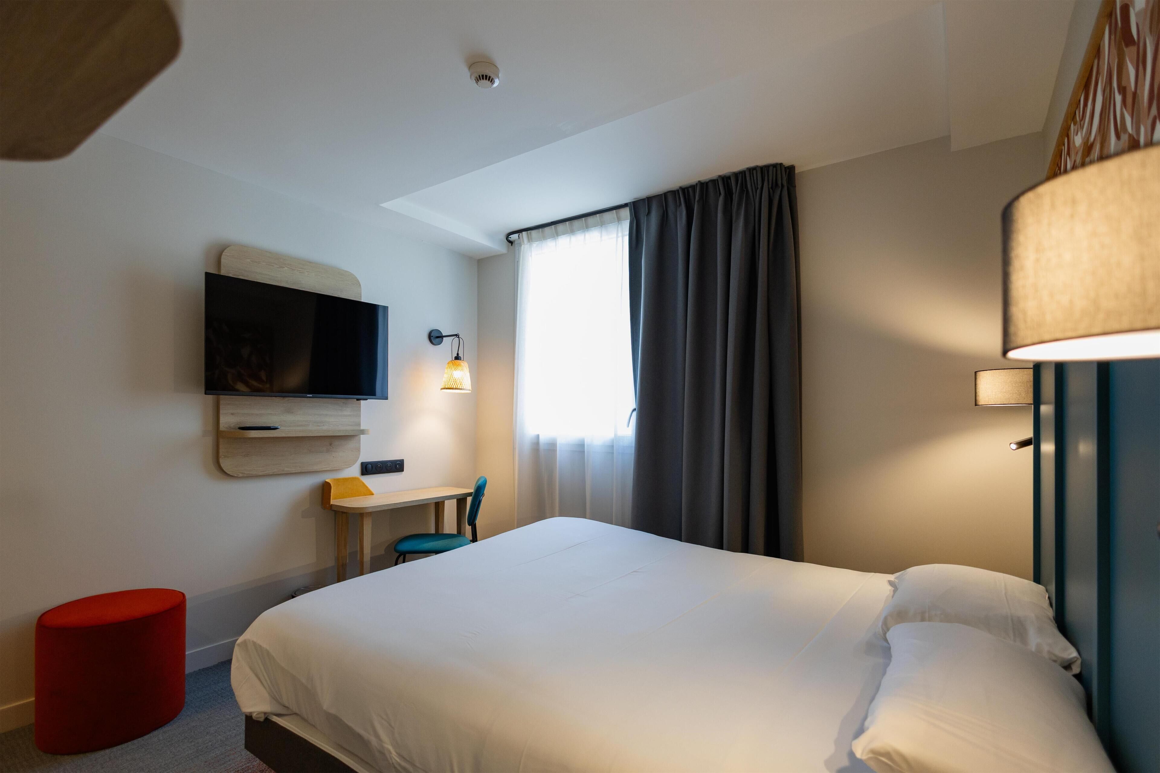 A hotel room with a bed, a desk, a chair, a television, and a window with curtains.