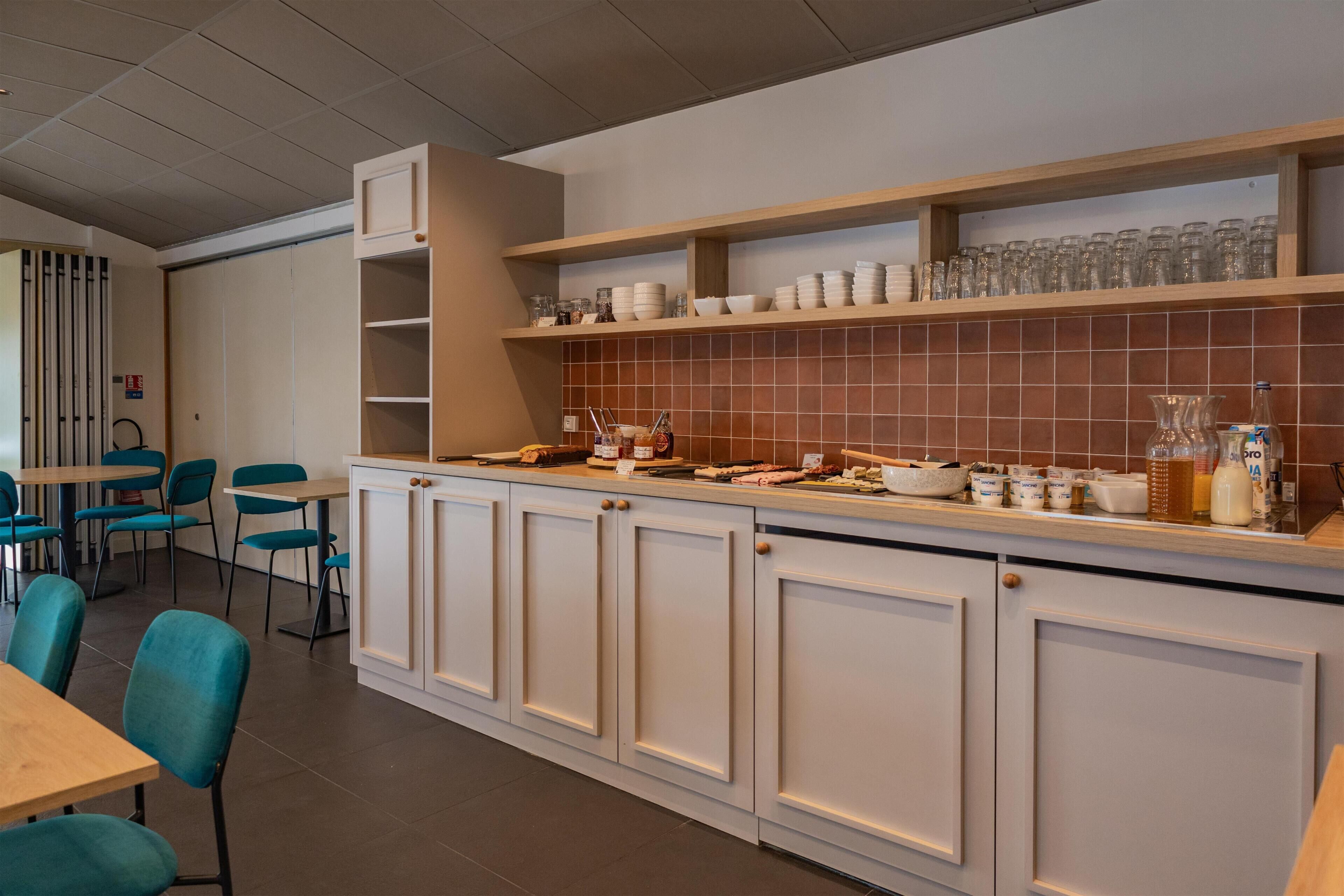 Una cocina moderna con armarios blancos, salpicadero de azulejos y una encimera con diversos utensilios de cocina. Hay sillas azules y una mesa en la zona de comedor.
