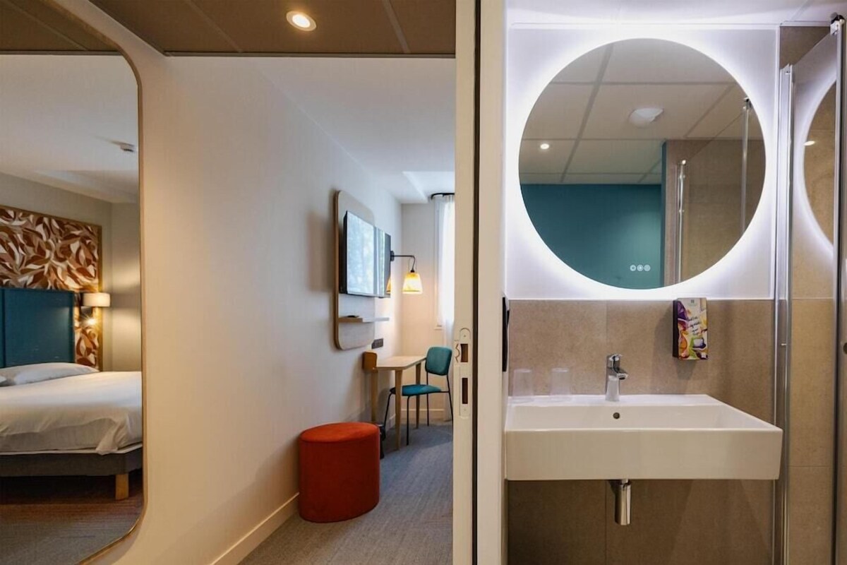 A modern hotel room with a bed, a round mirror, a small table, and a red ottoman.