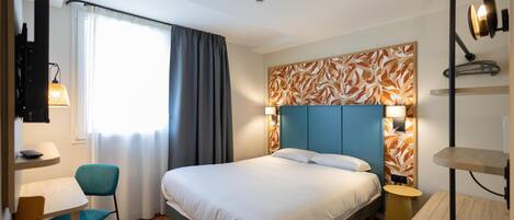 A hotel room with a bed, a desk, a chair, a lamp, and a wall-mounted TV.