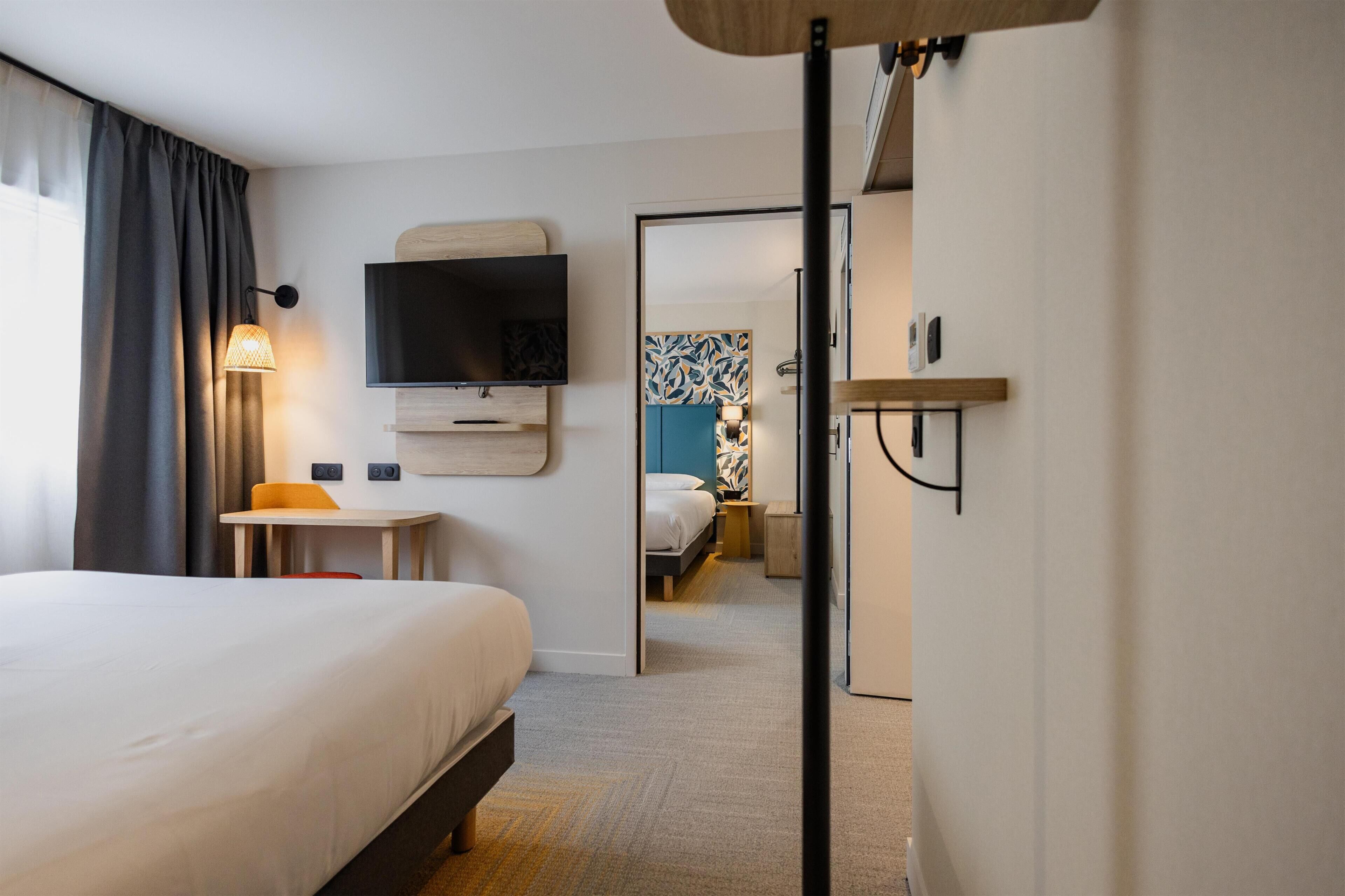 A hotel room with a bed, a desk, a television, a mirror, and a door leading to another room.