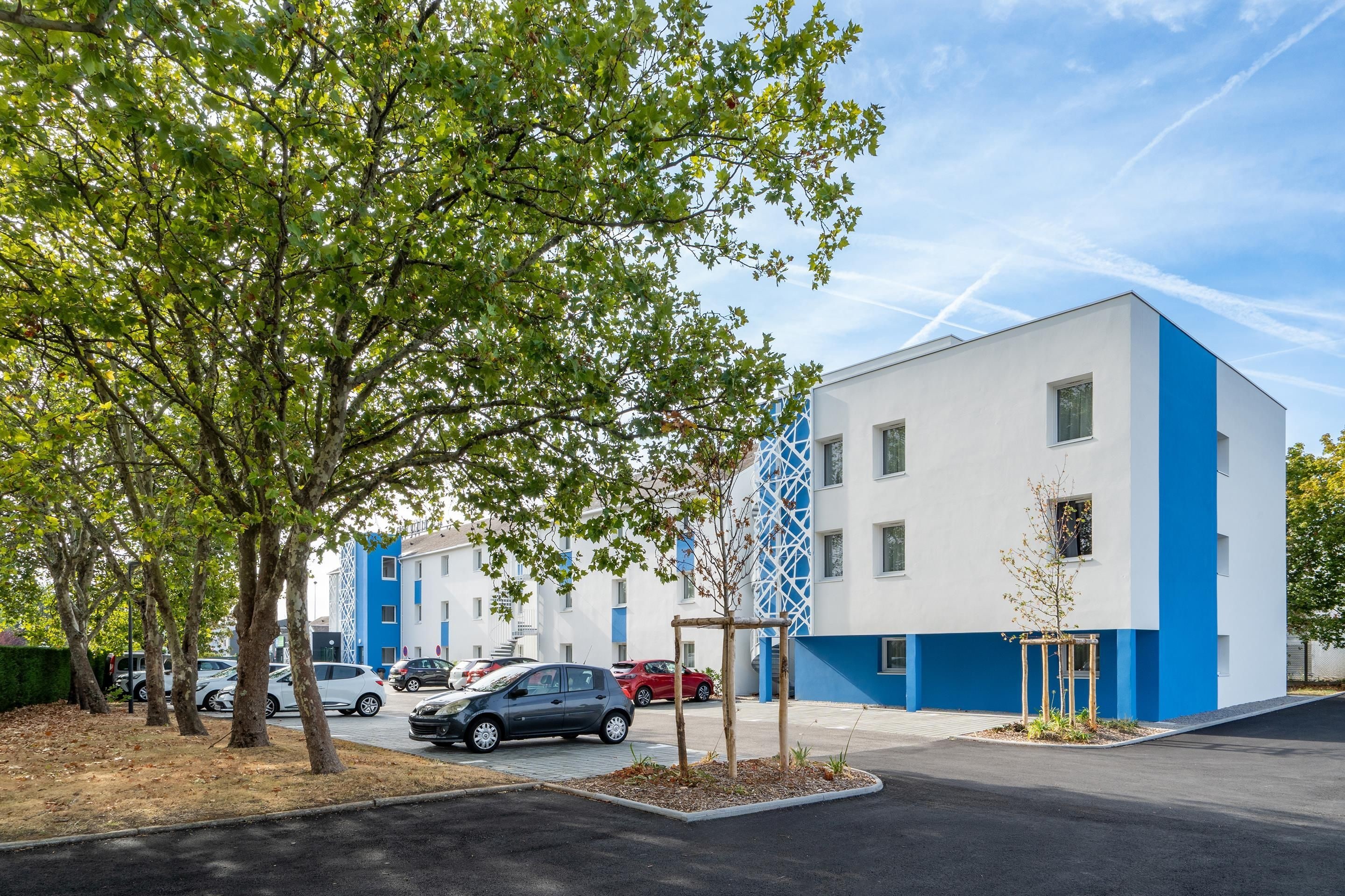 A modern building with a white and blue facade, surrounded by trees and parked cars.