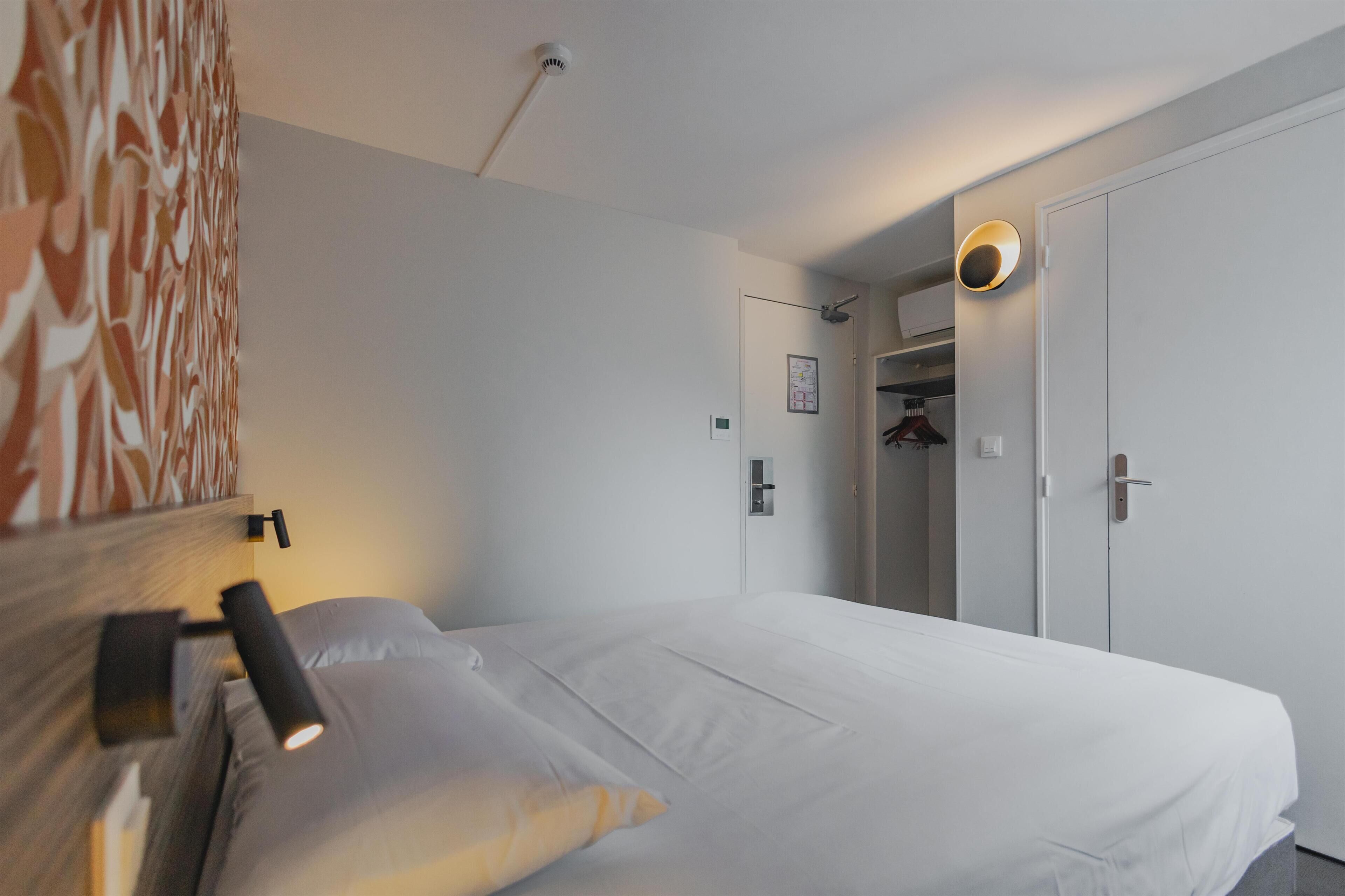 Habitación de hotel con cama, lámpara de noche, luz en la pared, una puerta y un armario.