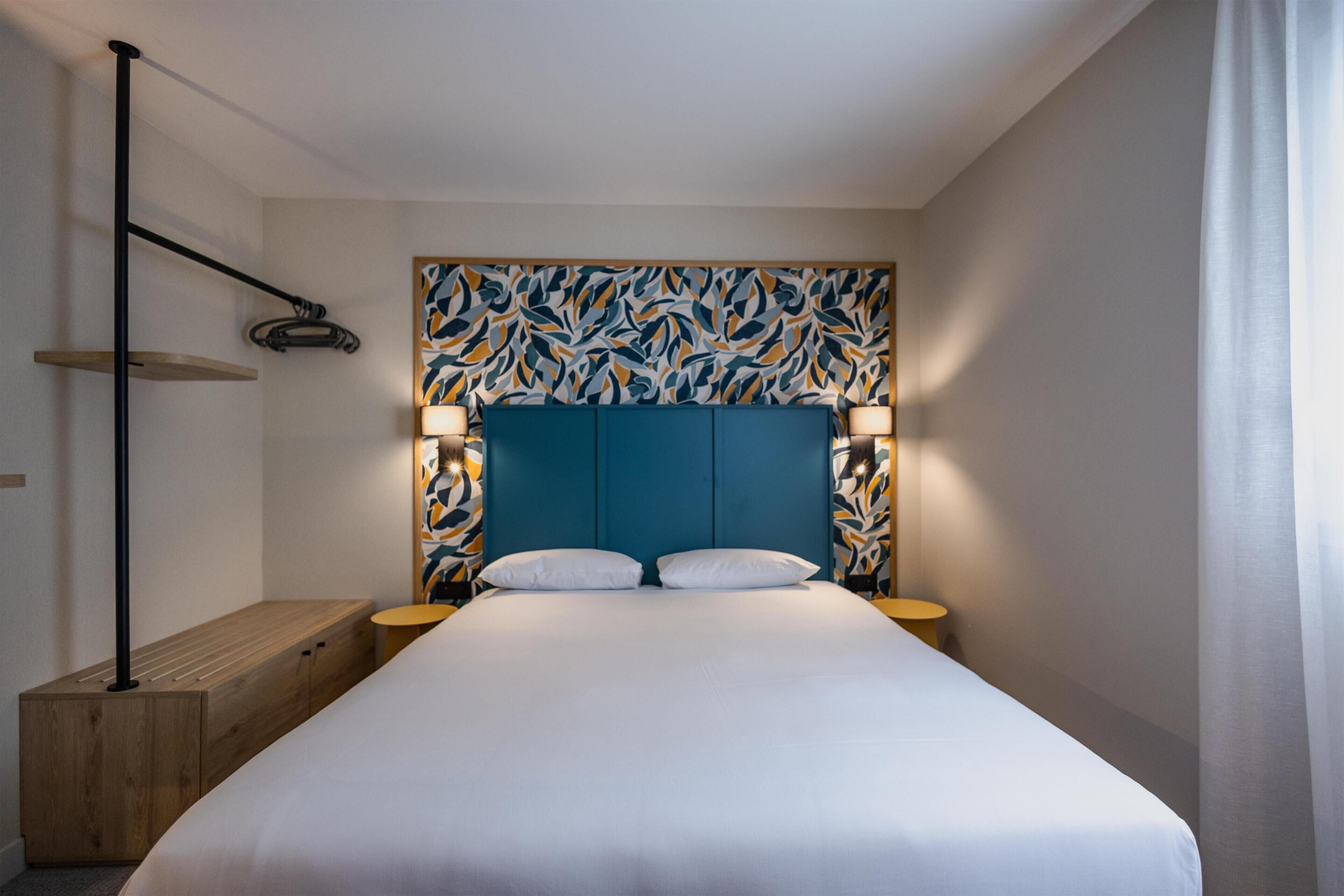 Une chambre d’hôtel moderne avec un grand lit, un banc en bois, deux tables de chevet avec des lampes et une tête de lit à motifs.