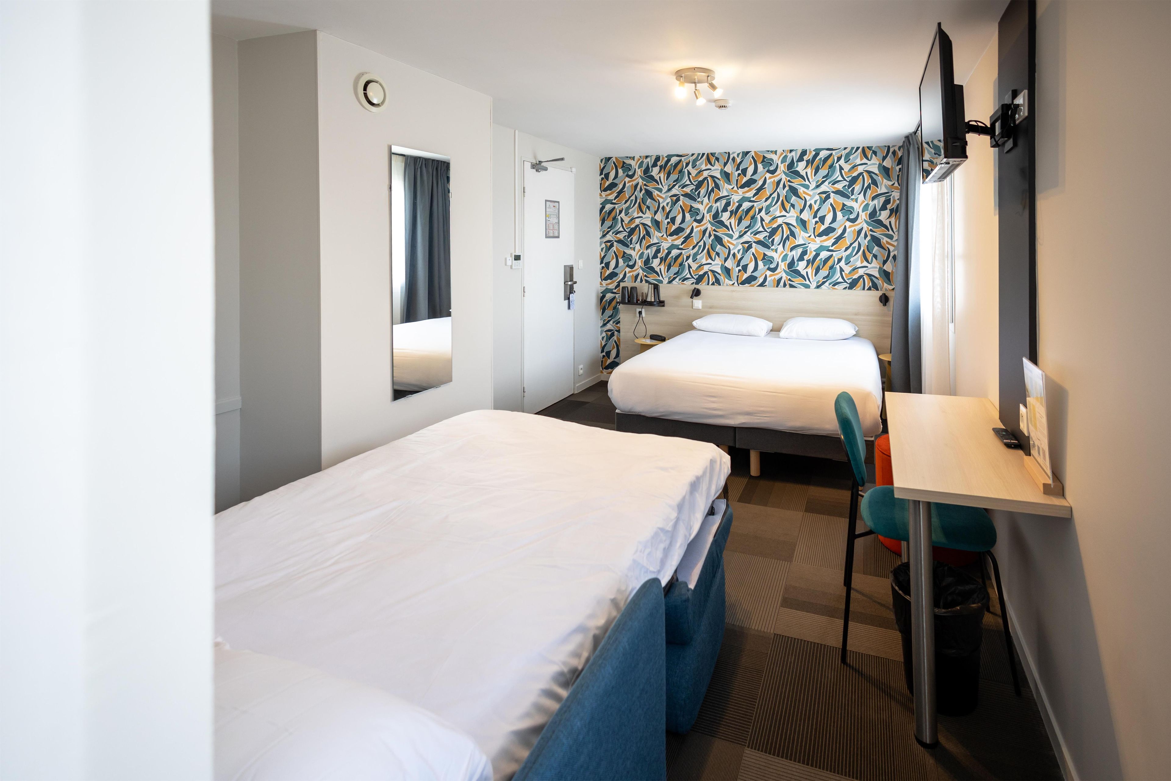 Habitación de hotel con dos camas, un escritorio, una silla, un espejo, un reloj y un papel tapiz estampado.