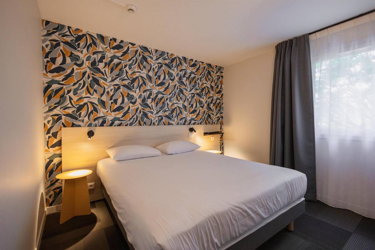 Ein Hotelzimmer mit einem großen Bett, einem Nachttisch, einer Wandleuchte und einem Fenster mit Vorhängen.