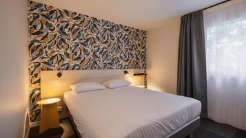 A hotel room with a large bed, a nightstand, a wall-mounted lamp, and a window with curtains.