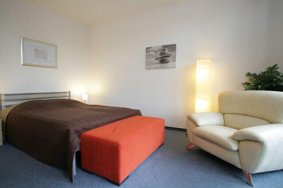 A bedroom with a bed, an ottoman, a chair, and a wall-mounted lamp.