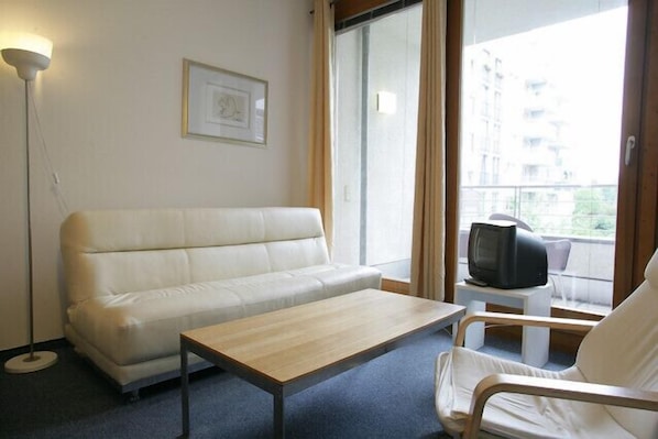 A room with a white sofa, a small wooden table, a television on a side table, and a large window with a view of buildings.