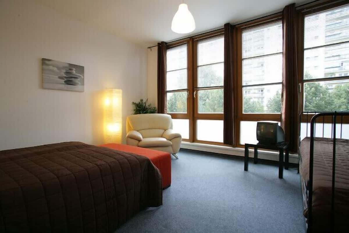 A room with two beds, a chair, a TV, and large windows.