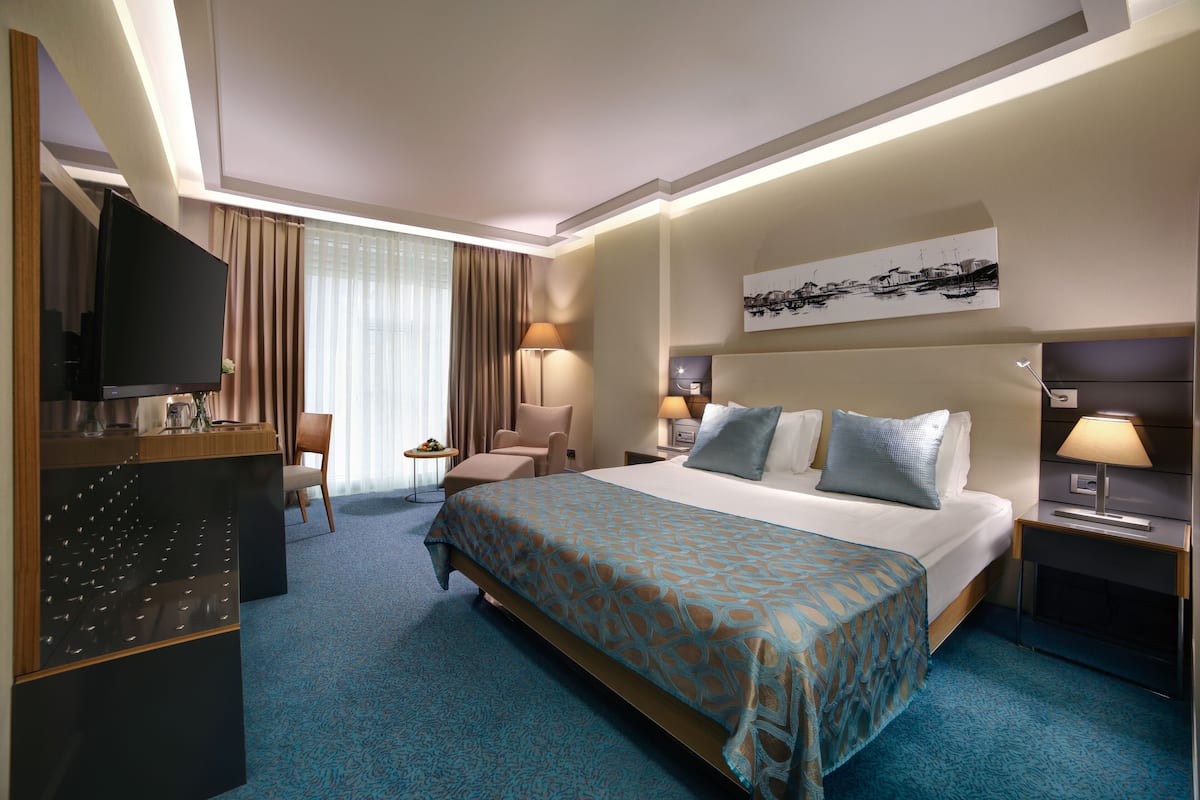 A hotel room with a large bed, a desk with a TV, a chair, a small table with a lamp, and a painting on the wall.