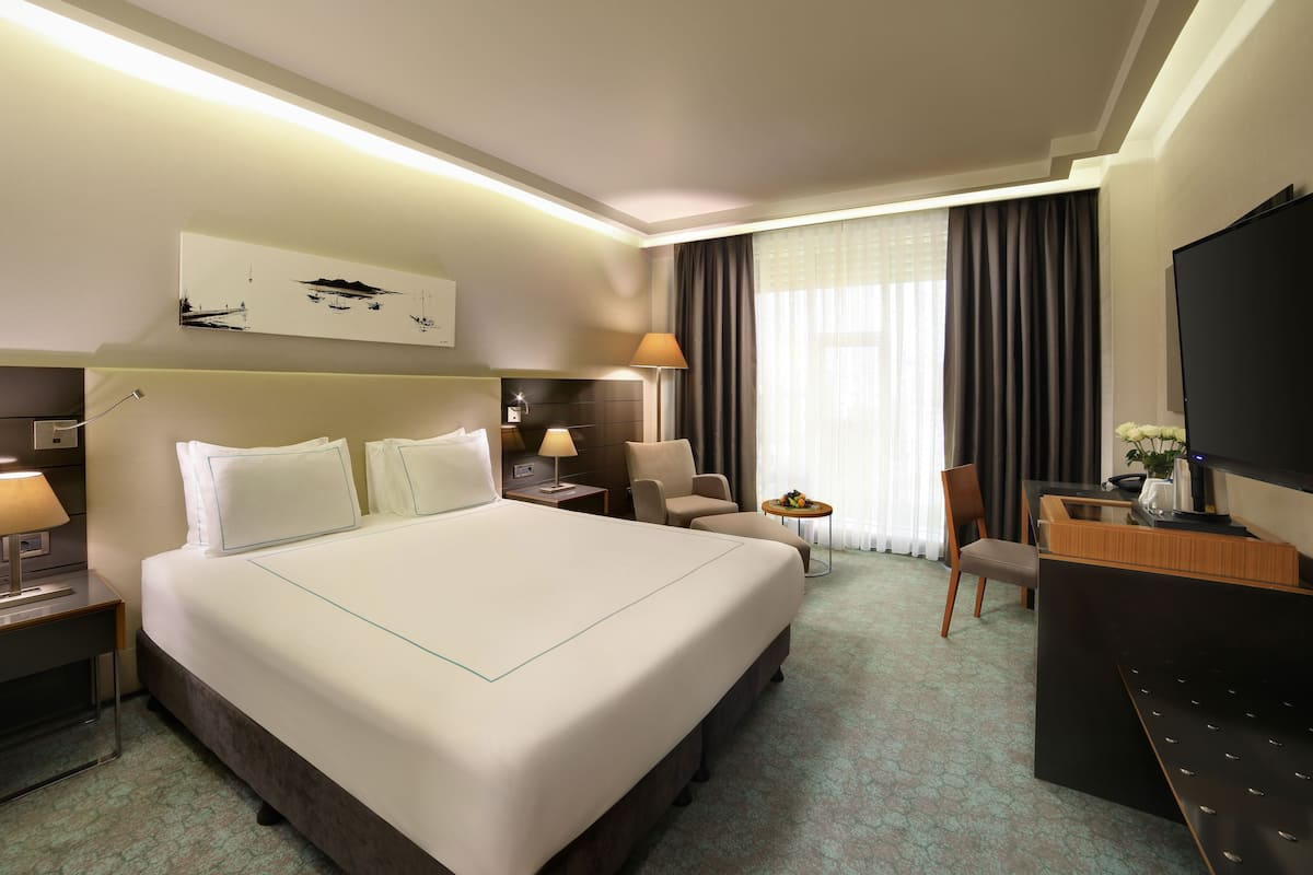 A hotel room with a large bed, bedside tables with lamps, a desk with a chair, a TV, and a painting on the wall.