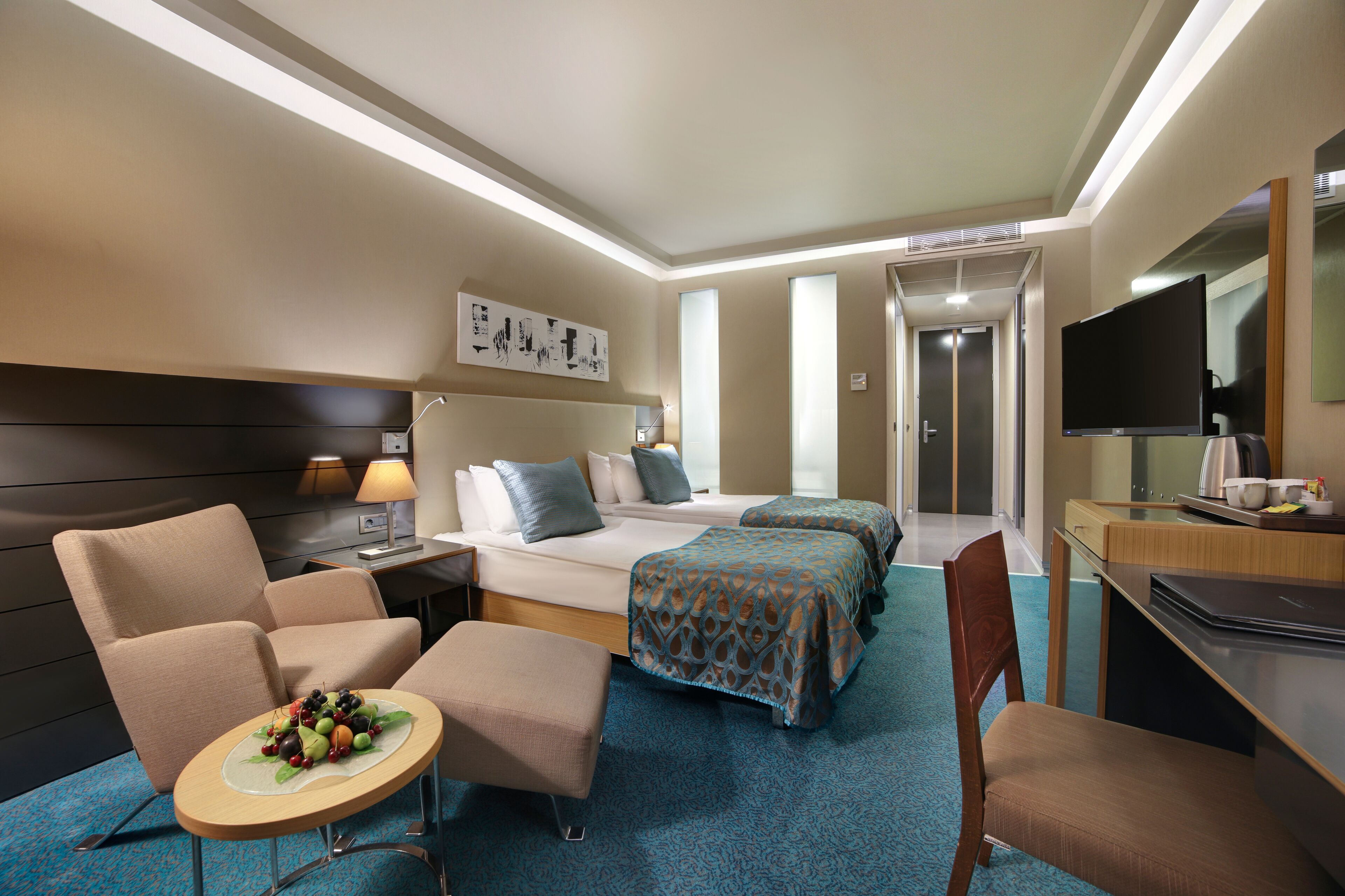 Een hotelkamer met een bed, twee fauteuils, een klein tafeltje met fruit, een bureau met een laptop en een televisie.