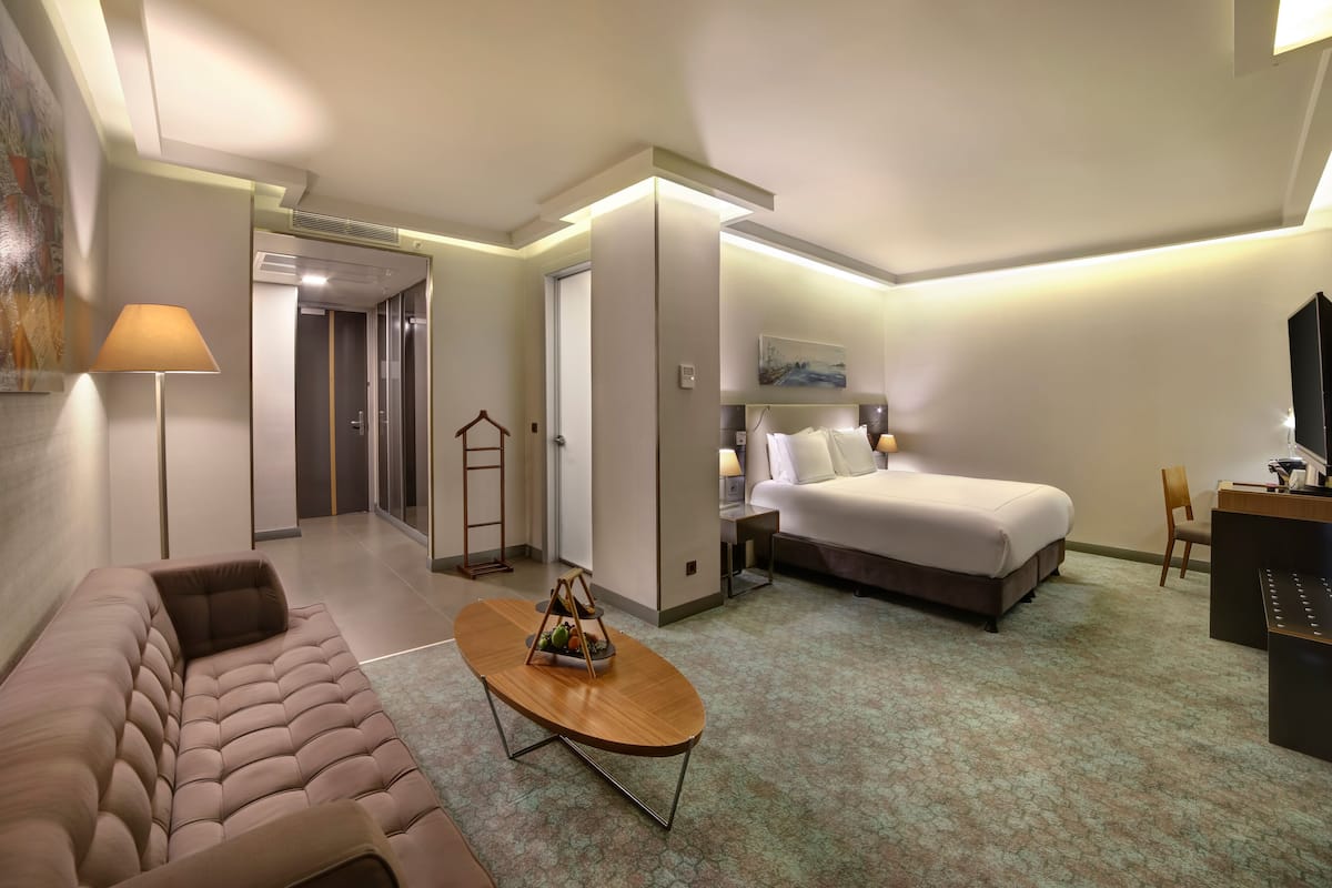 A hotel room with a sofa, a bed, a desk, a lamp, and a painting on the wall.