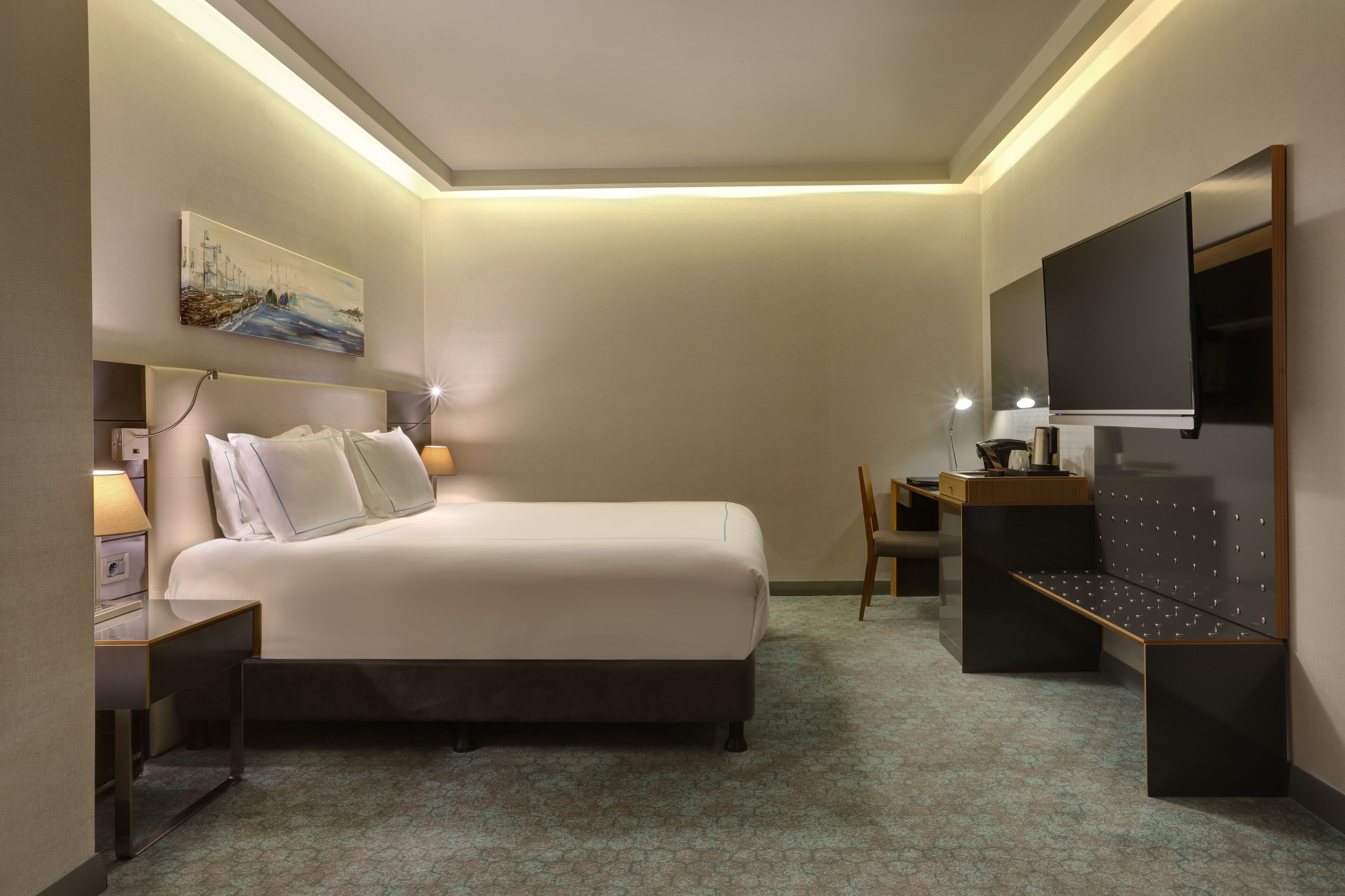 Quarto de hotel com cama, mesa de cabeceira, luminária, televisão e um quadro na parede.