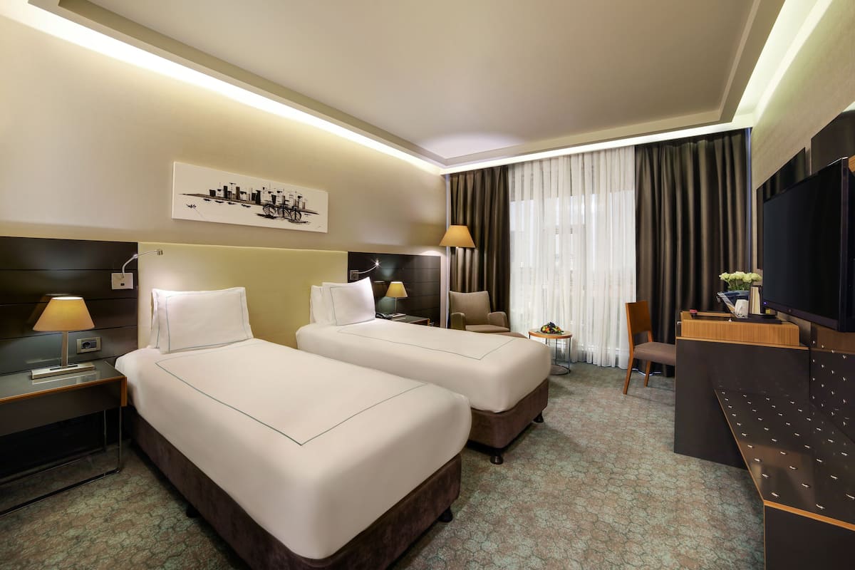 A hotel room with two beds, a desk, a TV, and a cityscape artwork on the wall.