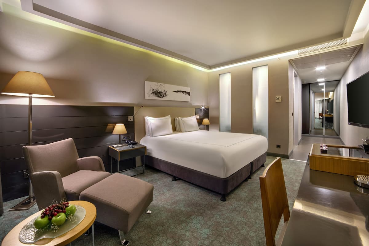 A hotel room with a large bed, a desk, a chair, a sofa, and a small table with fruit.