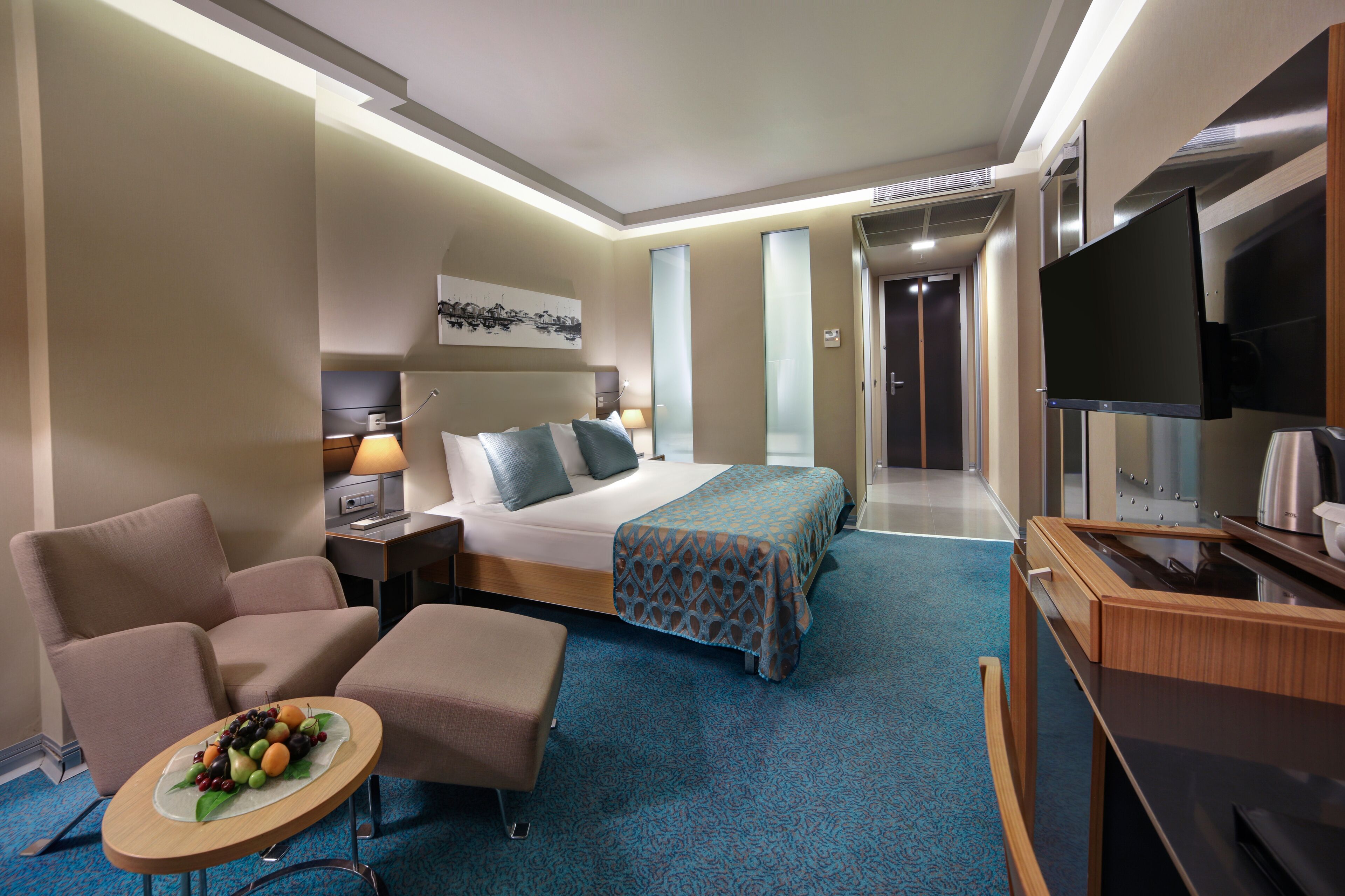 Ein Hotelzimmer mit Bett, Schreibtisch, Fernseher, Sessel und einem kleinen Tisch mit Obst.