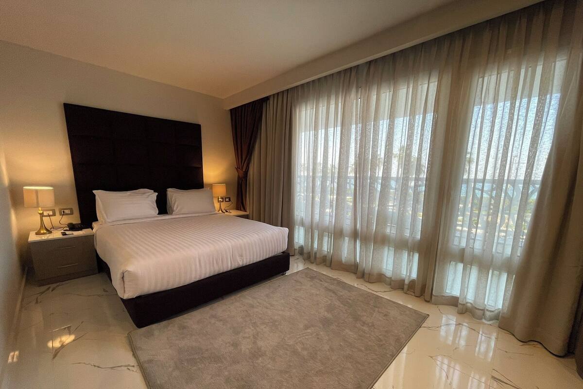 Una habitación de hotel con una cama grande, dos mesitas de noche, cabecera y ventanas del suelo al techo con cortinas translúcidas.