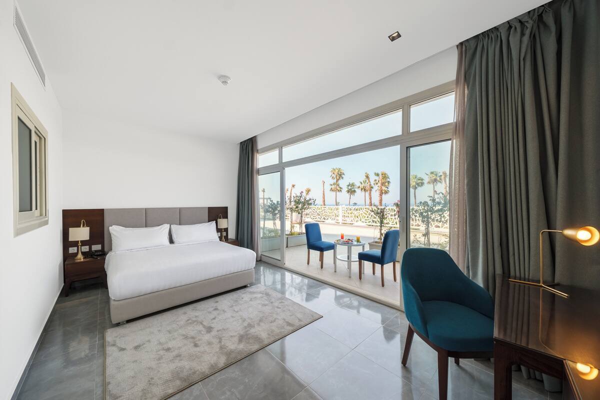 Una habitación de hotel moderna con una cama grande, un balcón con sillas y vista a palmeras.